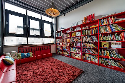 Der Leseraum der Picasso-Gruppe mit rotem Ecksofa, Teddy-Teppich und einer ganzen Wand voll Bücher